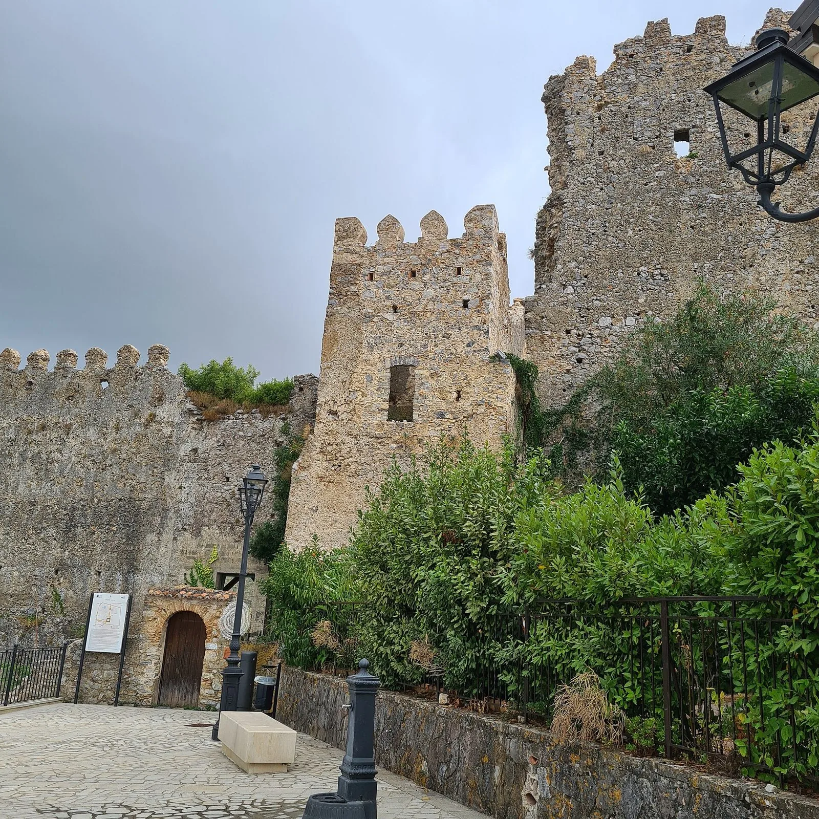 Mura merlate e torre in pietra del castello marchesale di Camerota, con una piazzetta lastricata e vegetazione ai piedi delle mura