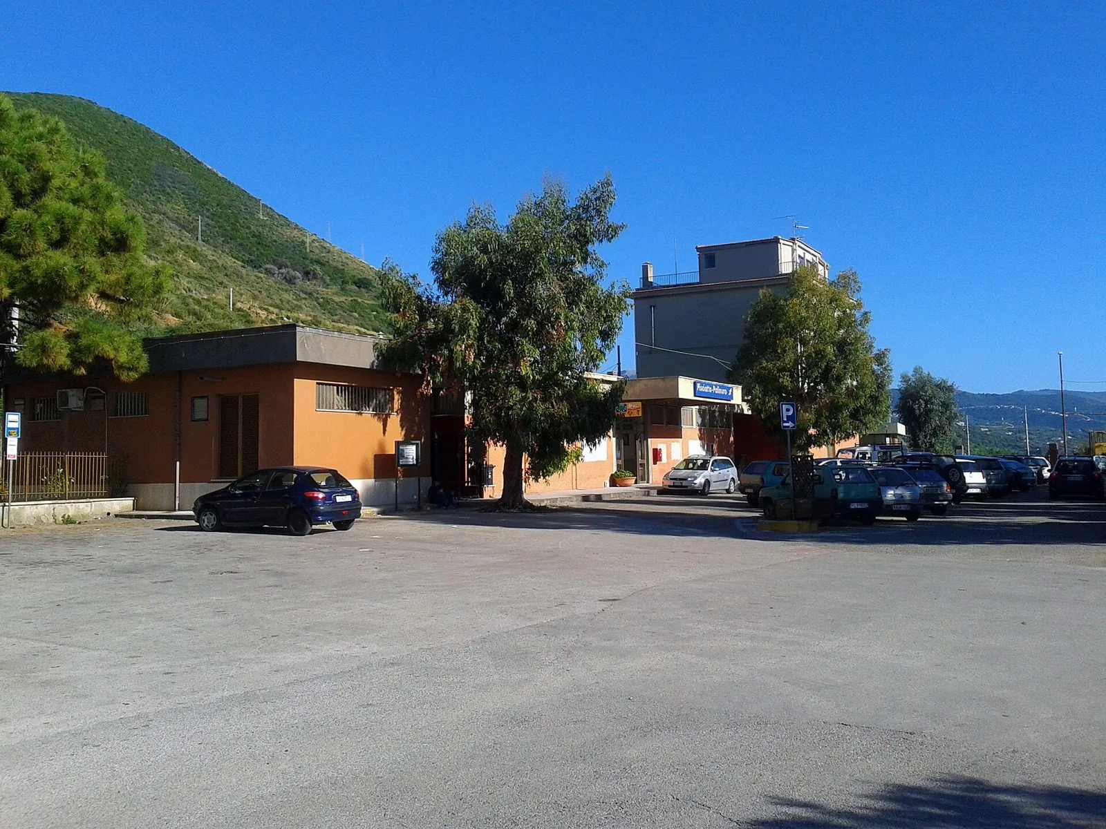 Il piazzale esterno della stazione ferroviaria di Pisciotta-Palinuro con l'edificio arancione, alcuni alberi e auto parcheggiate sotto una collina verde