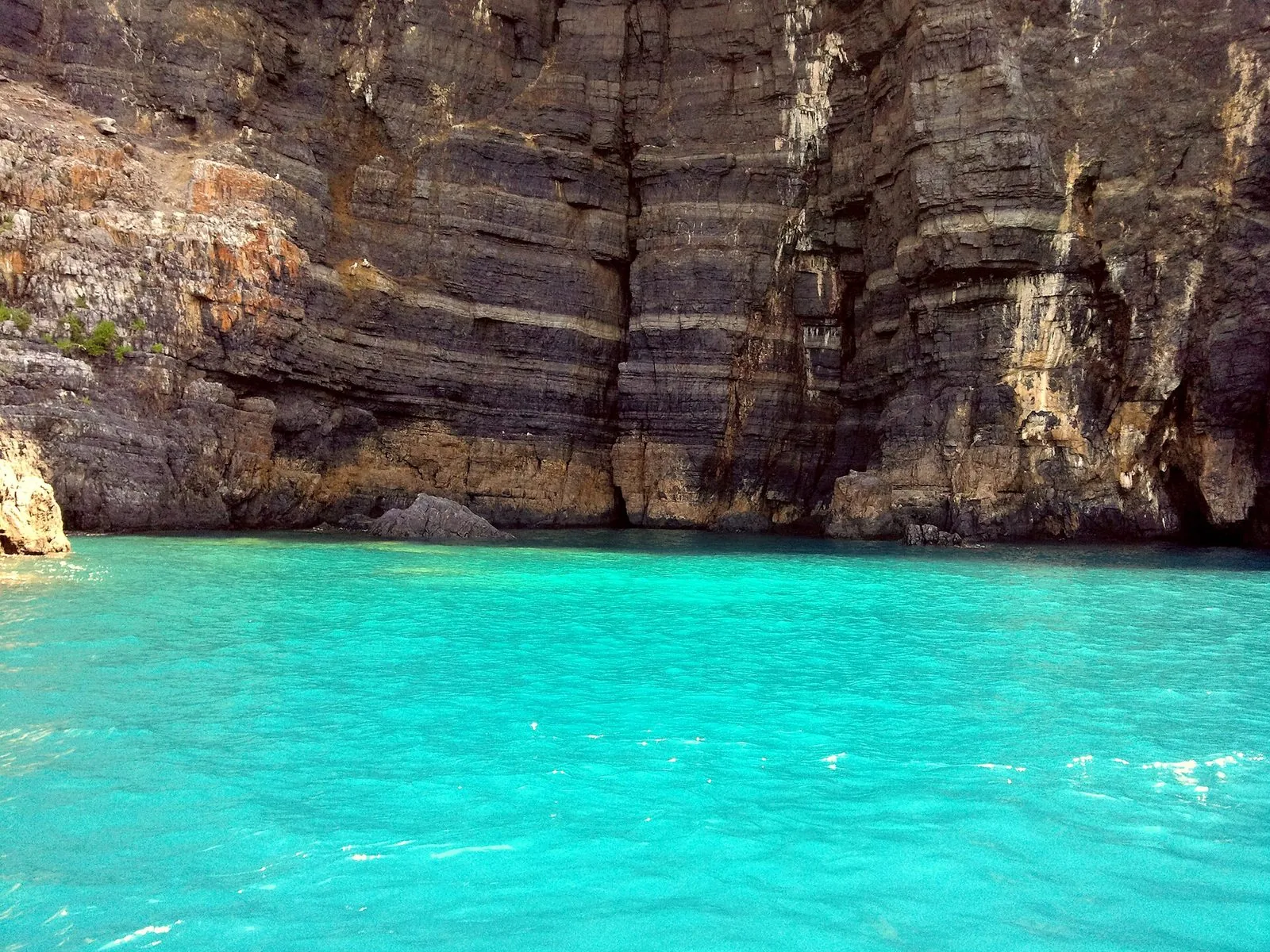Parete di roccia stratificata a picco su un mare turchese lungo la Costa degli Infreschi, vista da una barca