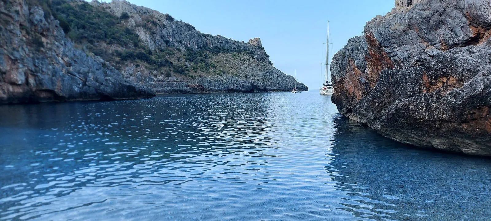 Insenatura di Cala Bianca con acqua limpida tra due pareti di roccia, barche a vela all'ancora e una torre di avvistamento sul promontorio